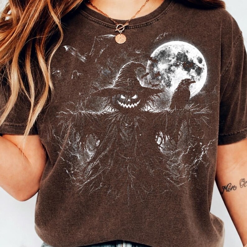 May include: A dark brown short-sleeve t-shirt featuring a white graphic print of a scarecrow with a jack-o'-lantern head wearing a wide-brimmed hat, standing beneath a full moon with a raven perched on its arm and bats flying in the background.