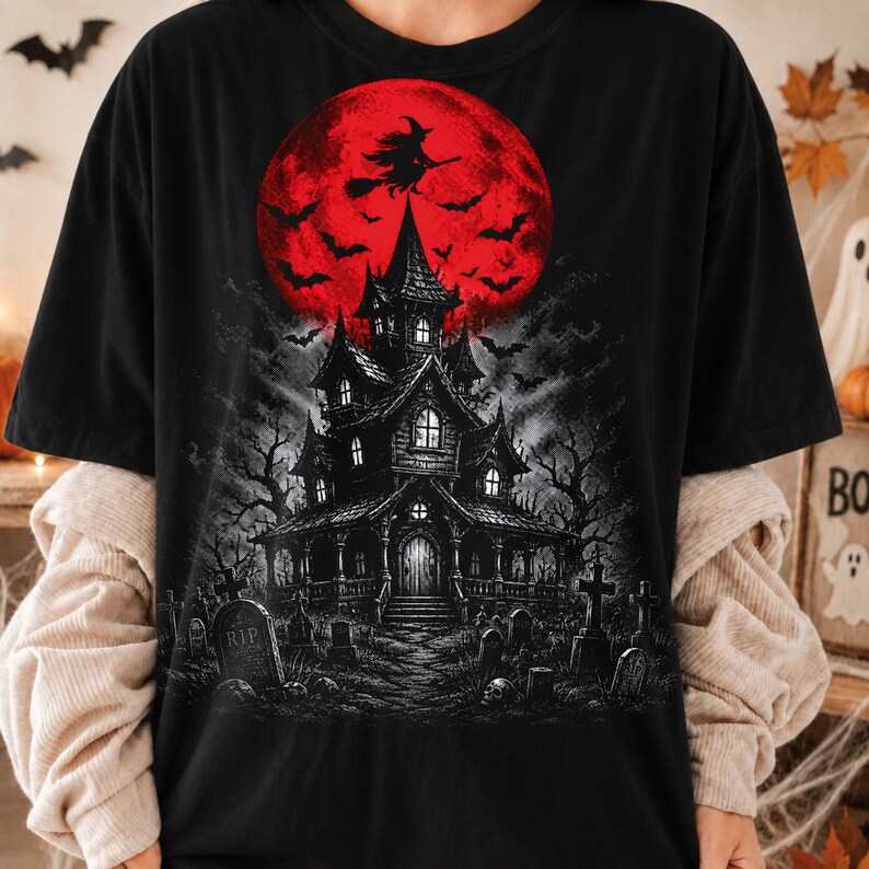 May include: A black t-shirt featuring a graphic print of a dark, multi-story haunted house set in a graveyard with tombstones. Above the house is a large, vibrant red full moon with a silhouette of a witch flying on a broomstick and bats.