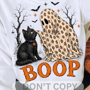 May include: A white t-shirt featuring a graphic of a black cat with a purple collar and a pumpkin charm, being touched on the nose by a ghost covered in a beige sheet with a brown flower pattern. The text BOOP is printed in large orange letters.