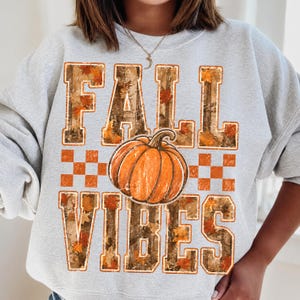 May include: A light gray crewneck sweatshirt featuring a graphic print with the text FALL VIBES in block letters. The letters have a camouflage and autumn leaf pattern. An orange pumpkin is centered between the words, flanked by orange checkered squares.