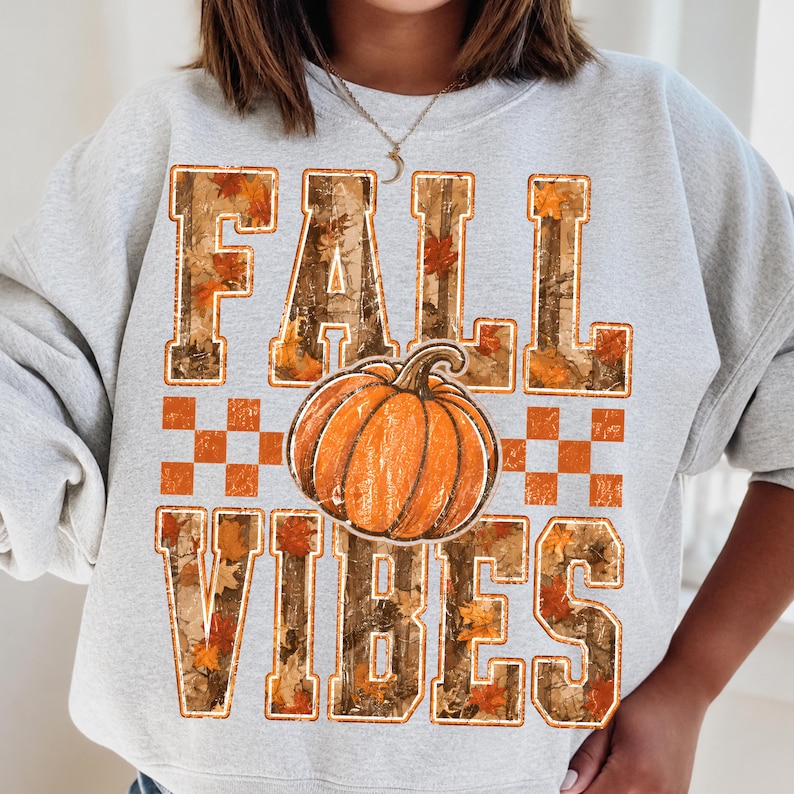 May include: A light gray crewneck sweatshirt featuring a graphic print with the text FALL VIBES in block letters. The letters have a camouflage and autumn leaf pattern. An orange pumpkin is centered between the words, flanked by orange checkered squares.