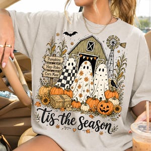 May include: A light gray t-shirt featuring a graphic print of three ghosts in front of a barn, pumpkins, corn stalks, a windmill, and a signpost labeled Pumpkins, Hay-Rides, and Corn Maze, with the text Tis the season written at the bottom.