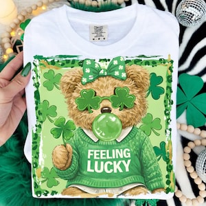 May include: White t-shirt featuring a cartoon teddy bear with shamrock glasses and a green sweater. The bear is blowing a green bubble and holding a shamrock. The shirt has the words "Feeling Lucky" in white.