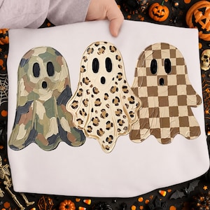 May include: A white t-shirt featuring a graphic print of three ghosts arranged side by side. The first ghost has a camouflage pattern, the second has a leopard print, and the third has a brown and white checkerboard pattern. Each ghost has black eyes.