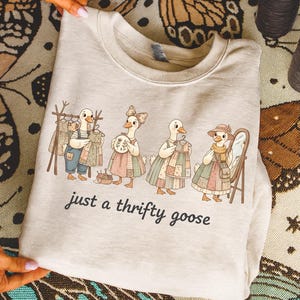 Może przedstawiać: Jasna beżowa bluza z zabawną ilustracją czterech gęsi w patchworkowych sukienkach, z lustrem i wieszakiem na ubrania. Tekst "just a thrifty goose" jest wydrukowany poniżej.