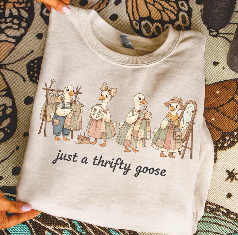 Może przedstawiać: Jasna beżowa bluza z zabawną ilustracją czterech gęsi w patchworkowych sukienkach, z lustrem i wieszakiem na ubrania. Tekst "just a thrifty goose" jest wydrukowany poniżej.