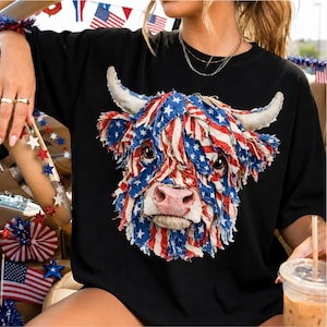 Pode incluir: Camiseta preta com um gráfico detalhado da cabeça de uma vaca Highland. O pelo da vaca é desenhado com um padrão de listras vermelhas e brancas e estrelas azuis, que se assemelha à bandeira americana. A vaca tem chifres brancos e um nariz rosa.