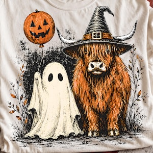 Puede incluir: Una camiseta color crema con una ilustración gráfica de una vaca Highland marrón de pelo largo con un sombrero de bruja negro con cuernos, junto a un fantasma blanco que sostiene un globo de calabaza naranja con rasgos negros.