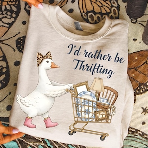 Puede incluir: Sudadera beige con un dibujo de un ganso con un lazo de leopardo y botas rosas, empujando un carrito de compras lleno de art&iacute;culos vintage. El texto dice: "I'd rather be Thrifting."