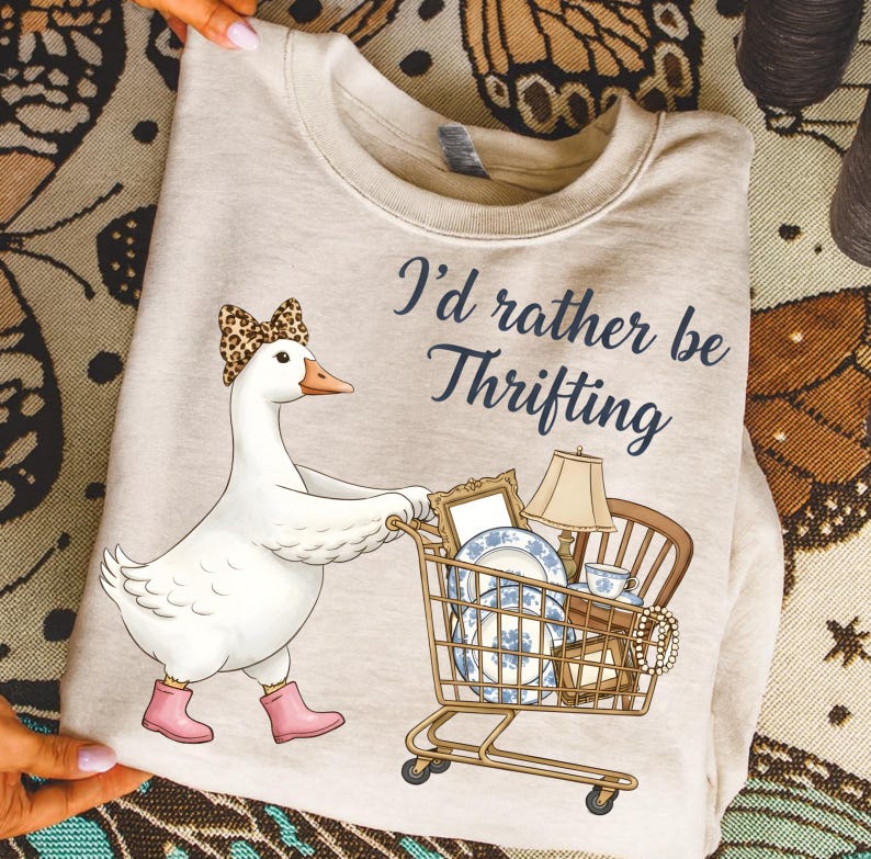 Puede incluir: Sudadera beige con un dibujo de un ganso con un lazo de leopardo y botas rosas, empujando un carrito de compras lleno de art&iacute;culos vintage. El texto dice: "I'd rather be Thrifting."