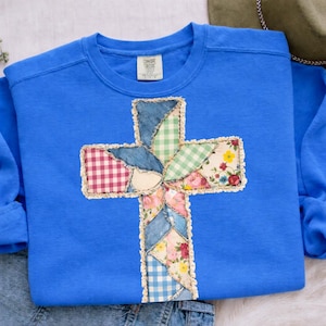 Könnte beinhalten: Blaues Sweatshirt mit einem Patchwork-Kreuz-Design. Das Kreuz besteht aus verschiedenen Stoffmustern, darunter Jeansstoff, Vichy-Karo und Blumenmuster. Das Sweatshirt hat einen Rundhalsausschnitt und lange Ärmel.