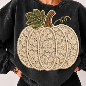 Peut inclure: Un sweat-shirt à col rond gris anthracite orné d'un grand motif de citrouille sur le devant. La citrouille est composée d'un tissu en dentelle florale crème avec une feuille brodée vert foncé et une tige brodée marron.