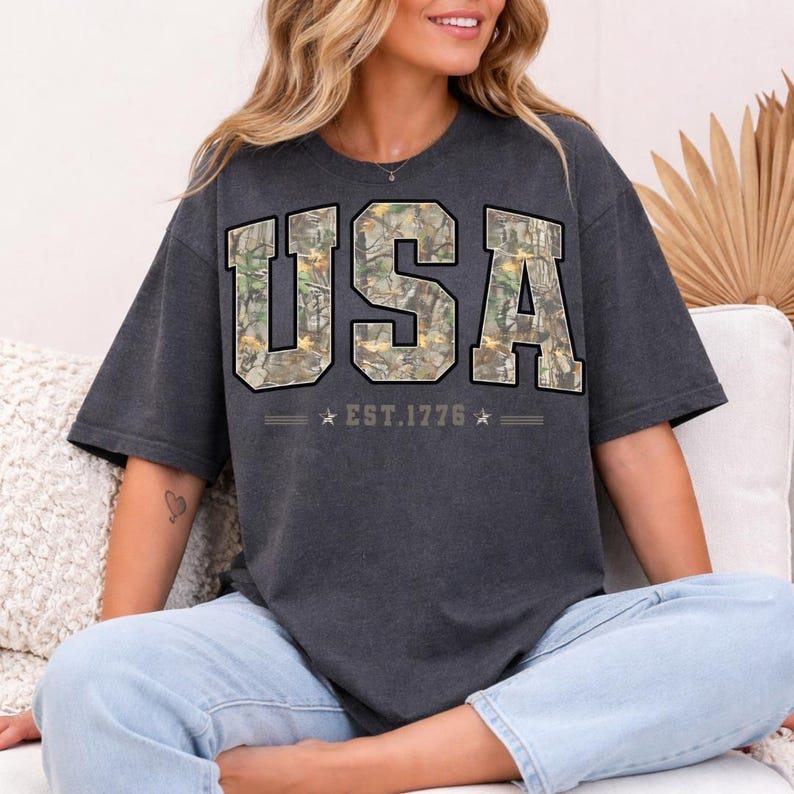 May include: Dark gray t-shirt with the word "USA" in large, block letters filled with a camouflage pattern. Below the word "USA" is the text "EST. 1776" with stars on each side. The shirt has short sleeves.