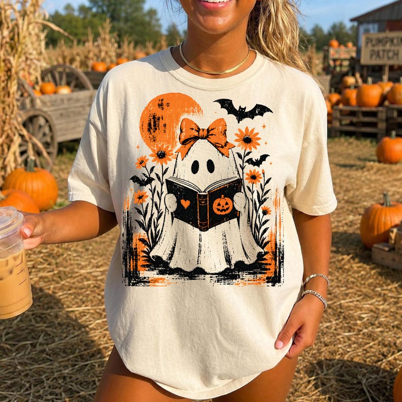 Pu&ograve; includere: Una t-shirt a maniche corte color crema con una stampa grafica di un fantasma che indossa un fiocco arancione, intento a leggere un libro con l'icona di una zucca. Il design include una luna arancione, pipistrelli neri e girasoli arancioni.