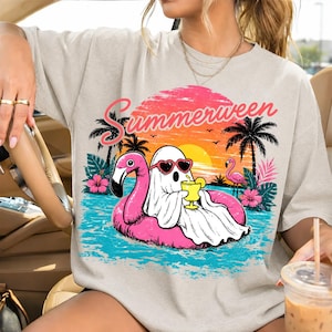 May include: Beige t-shirt with a colourful graphic. It shows a ghost in heart-shaped sunglasses on a pink flamingo float, with the word "Summerween", palm trees, and a sunset. The design evokes a summer holiday theme.