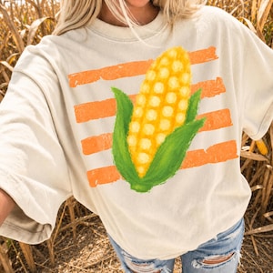 May include: A cream-colored t-shirt featuring a graphic of a yellow corn cob with green husks centered over horizontal, distressed orange stripes. The design has a textured, painted appearance on the front of the short-sleeved garment.