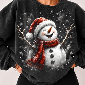 Puede incluir: Una sudadera negra de cuello redondo con lavado mineral que presenta un estampado gráfico de un muñeco de nieve sonriente con un gorro de Papá Noel rojo y una bufanda roja a juego. Tiene brazos de ramitas y tres botones negros en el torso.