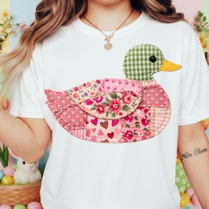 K&ouml;nnte beinhalten: Wei&szlig;es T-Shirt mit einem Patchwork-Enten-Design. Die Ente hat einen gr&uuml;nen Vichy-Kopf, einen gelben Schnabel und einen K&ouml;rper aus rosa Blumen- und Polka-Dot-Stoffen. Das Design ist mittig auf dem Shirt platziert.