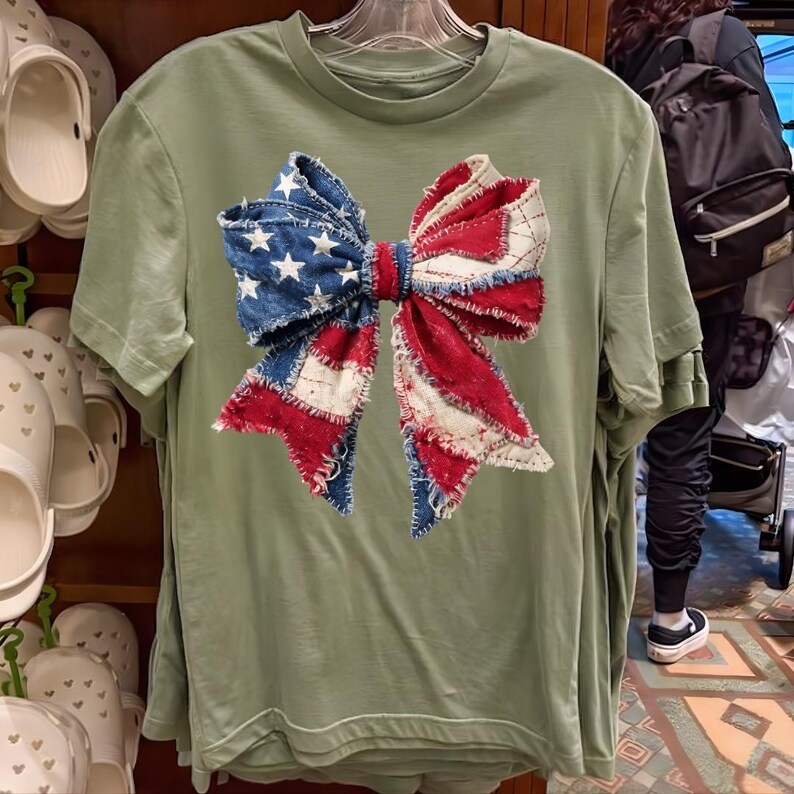 K&ouml;nnte beinhalten: Ein hellgr&uuml;nes T-Shirt mit einem gro&szlig;en Schleifendesign. Die Schleife besteht aus Jeansstoff sowie rotem und wei&szlig;em Stoff und &auml;hnelt der amerikanischen Flagge. Die Schleife hat ein verwaschenes Patchwork-Aussehen.