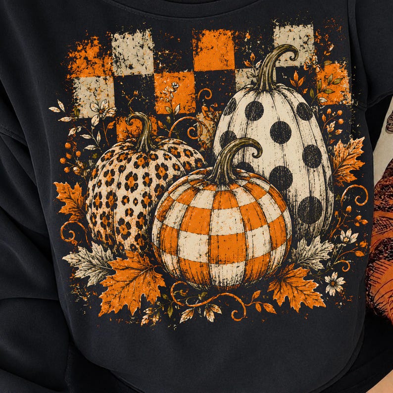 May include: A black sweatshirt featuring a graphic print of three pumpkins with different patterns: one with leopard print, one with black polka dots, and one with an orange and white checkerboard pattern, surrounded by orange autumn leaves.