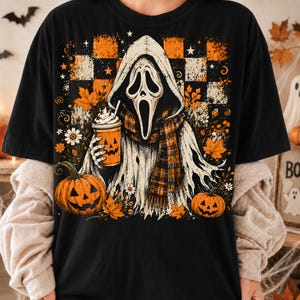 May include: A black t-shirt featuring a graphic of a hooded ghost figure wearing a plaid scarf and holding a pumpkin-faced cup. The design includes orange and white checkered patterns, autumn leaves, daisies, and two jack-o'-lantern pumpkins.