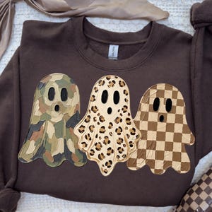 May include: A dark brown crewneck sweatshirt featuring three ghost graphics. The left ghost has a green and brown camouflage pattern, the middle ghost has a tan and black leopard print, and the right ghost has a brown and tan checkerboard pattern.