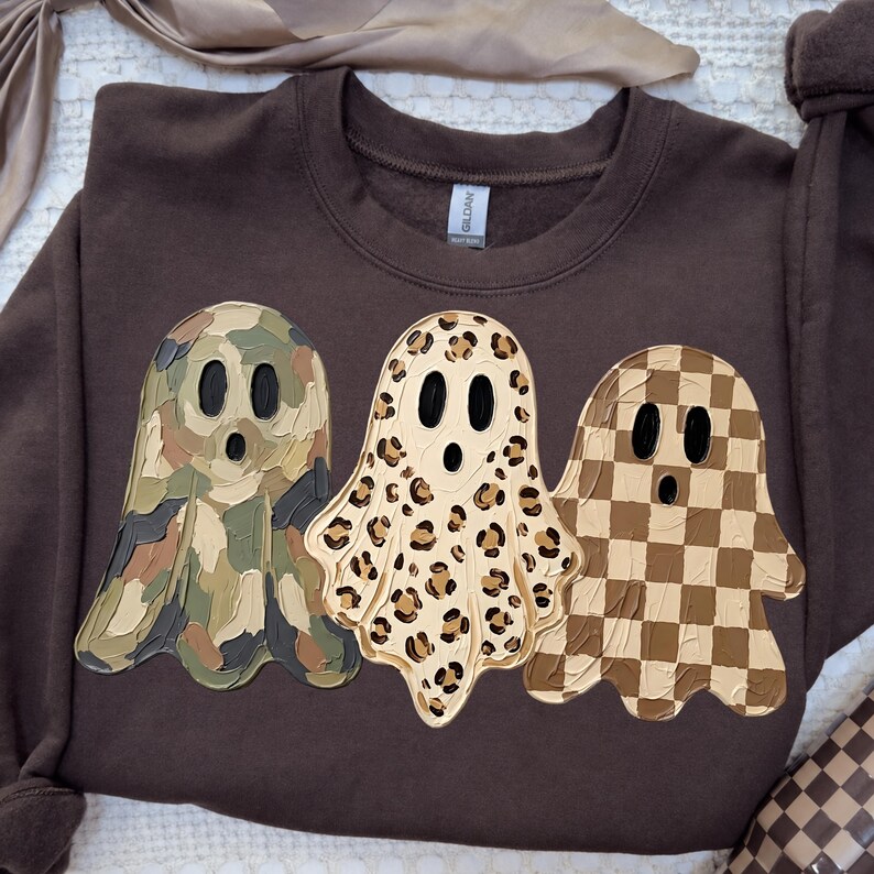 May include: A dark brown crewneck sweatshirt featuring three ghost graphics. The left ghost has a green and brown camouflage pattern, the middle ghost has a tan and black leopard print, and the right ghost has a brown and tan checkerboard pattern.