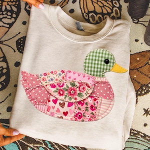 May include: A pale grey sweatshirt with a patchwork duck appliqué. The duck has a green gingham head, a yellow beak, and a body made of pink floral and polka dot fabrics. The sweatshirt is folded, showcasing the duck design.