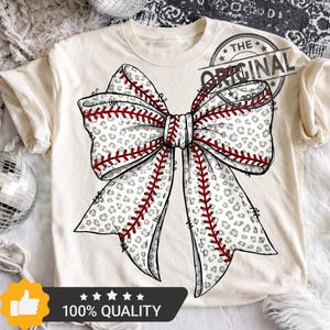 May include: Cream-colored t-shirt featuring a large bow design. The bow has a baseball stitch pattern with red thread and a leopard print background. The text "THE ORIGINAL" is stamped on the upper right. The bottom of the shirt has a "100% QUALITY" graphic.