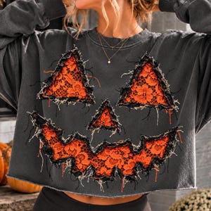 Könnte beinhalten: Ein anthrazitfarbenes Langarm-Sweatshirt mit einem großen Kürbisgesicht-Motiv. Die Augen, die Nase und der gezackte Mund sind mit leuchtend orangefarbenem Spitzenstoff gefüllt und mit ausgefransten, abgenutzten Kanten versehen.