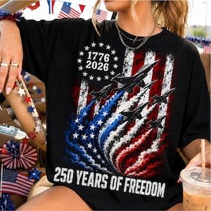 Pu&ograve; includere: T-shirt nera con un design patriottico raffigurante la bandiera americana, jet e il testo "1776 2026" e "250 YEARS OF FREEDOM". Il design incorpora i colori rosso, bianco e blu.