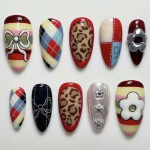 Könnte beinhalten: Eine Sammlung von zehn künstlichen Nägeln mit verschiedenen Designs. Die Designs umfassen Argyle-Muster, Schleifen, Leopardenmuster und florale Akzente. Farben sind Rot, Weiß, Blau, Schwarz und Silber. Die Nägel sind oval geformt.