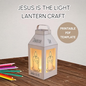 Op de afbeelding: Een witte papieren lantaarn met de tekst Jesus Is en John 8:12 en kaarsillustraties op de zijkanten. De lantaarn heeft een handvat en is van binnenuit verlicht. Een set kleurpotloden ligt op het oppervlak naast de lantaarn.