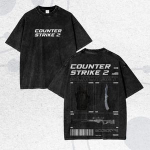 May include: Black t-shirt with the text "COUNTER STRIKE 2" in white on the front. The back of the shirt features the text "COUNTER STRIKE 2 BLACK SET" with graphic illustrations of a glove, knife, and rifle. The shirt has a vintage, washed appearance.
