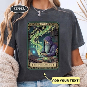 May include: Dark grey t-shirt with a tarot card design. The card shows a wizard in a purple hood using a laptop with a glowing Apple logo. The card is framed in green and gold with the text "THE PROGRAMMER".