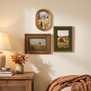 May include: Three framed wall art pieces featuring landscape paintings. One oval gold frame shows a person and dog in a field. A rectangular brown beaded frame displays two dogs in grass. A rectangular green velvet frame shows haystacks in a field.