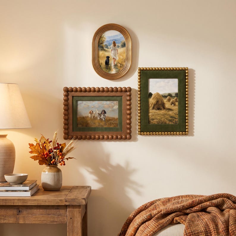 May include: Three framed wall art pieces featuring landscape paintings. One oval gold frame shows a person and dog in a field. A rectangular brown beaded frame displays two dogs in grass. A rectangular green velvet frame shows haystacks in a field.