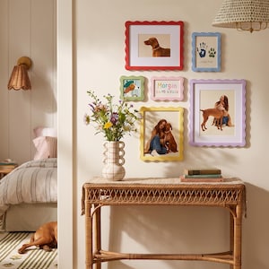 May include: A collection of six colorful, scalloped-edge picture frames arranged on a wall above a rattan console table. The frames are red, light blue, green, pink, yellow, and lavender, displaying various photographs and a handprint art piece.