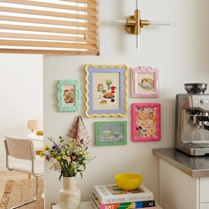 Puede incluir: Una colección de obras de arte enmarcadas con marcos decorativos coloridos, que incluyen un marco verde claro con una ilustración de hongo, un marco amarillo y morado con una ilustración de comida y un marco rosa con una ilustración de taza de té. También se ve un jarrón de flores y una cafetera.