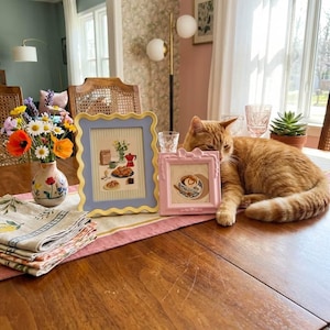Peut inclure: Une table en bois présente un arrangement floral dans un vase peint, deux illustrations encadrées et une serviette en lin pliée. Un chat roux se repose près des cadres. Les cadres sont de couleur bleu clair et rose.