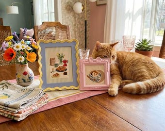 May include: A wooden table displays a floral arrangement in a painted vase, two framed illustrations, and a folded linen napkin. A ginger cat rests on the table near the frames. The frames are a light blue and pink color.