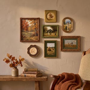 May include: A gallery wall arrangement featuring six framed landscape and animal paintings in various shapes and sizes, including rectangular, oval, and scalloped frames with beaded details, displayed above a wooden bench with books and a vase.