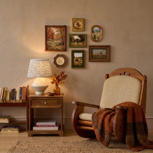 May include: A wooden rocking chair with a cream-colored cushion and a thick, rust-colored knit throw blanket. Beside it, a wooden side table holds a white lamp with a blue floral shade, a small amber vase with autumn leaves, and books.