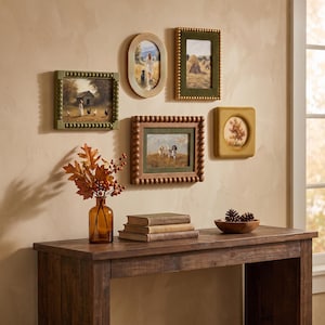 Puede incluir: Una colección de cinco cuadros de pared con escenas rurales, expuestos sobre una mesa consola de madera. El conjunto incluye varios estilos de marcos, como diseños ovalados y con cuentas, junto a un jarrón de cristal con follaje otoñal.