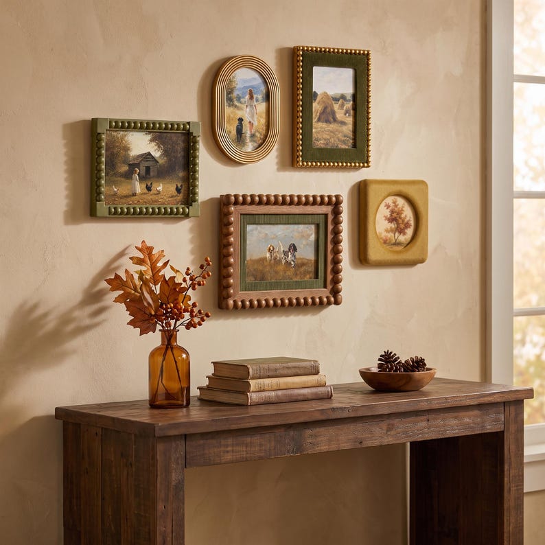 May include: A collection of five framed wall art pieces featuring rural scenes, displayed above a wooden console table. The arrangement includes various frame styles, including beaded and oval designs, alongside a glass vase with autumn foliage.