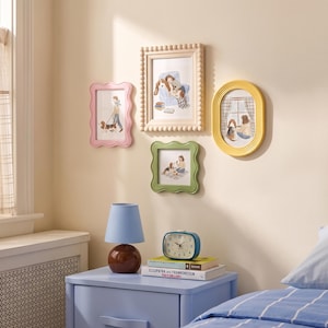 May include: A collection of four wall-mounted picture frames in pink, cream, green, and yellow, displaying illustrations of people and dogs. Below, a blue lamp with a brown base sits on a blue nightstand next to a teal alarm clock and books.