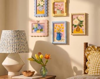 Peut inclure: Cinq cadres muraux rectangulaires présentant des natures mortes florales, avec des vases contenant des fleurs blanches, roses, bleues et jaunes. Les cadres ont des bords festonnés ou perlés en bleu, jaune et rose. Une lampe et un vase sont visibles.