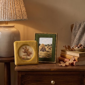 May include: A square mustard yellow velvet picture frame with a circular opening and a rectangular dark green velvet picture frame with a gold beaded border. The square frame displays a tree illustration, and the rectangular frame shows haystacks.