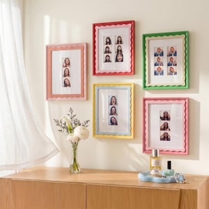 May include: Five rectangular picture frames with scalloped edges in pink, red, green, yellow, and rose colors, each containing vertical photo strips. The frames are arranged on a wall above a wooden surface with a glass vase of white flowers.