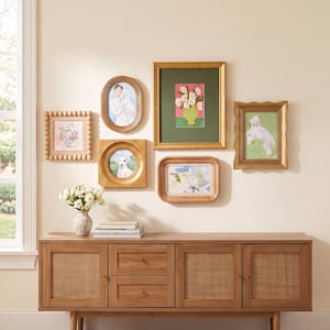 May include: A wooden sideboard with two drawers and two cane-paneled doors, topped with a white vase of flowers and a gallery wall of six framed artworks, including portraits of dogs and floral paintings in various gold and wooden frames.